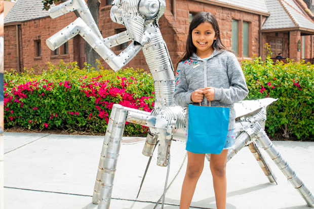 A girl is smiling standing in front of the Praying Mantis art piece holding a blue bag. 